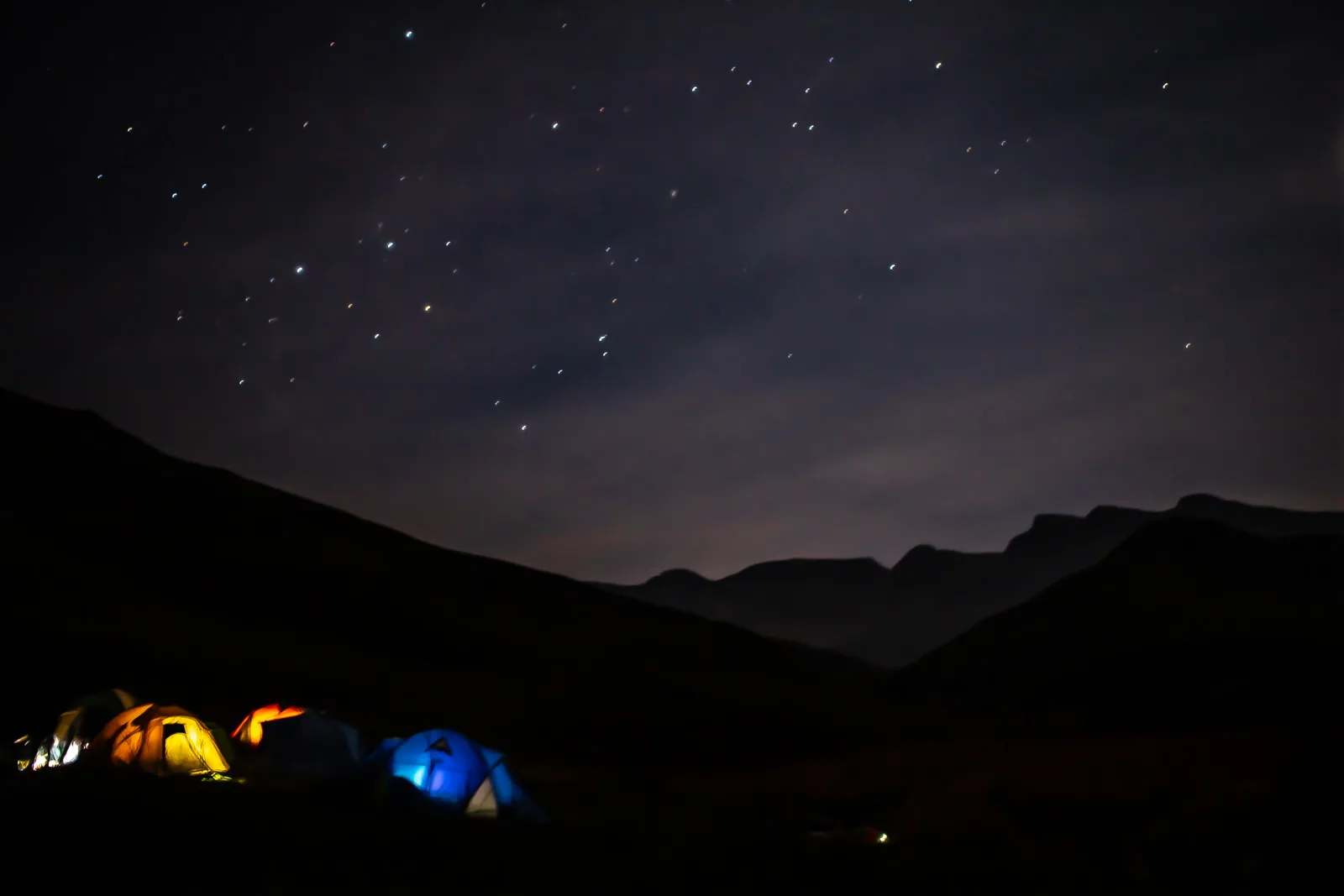 Illuminated tents beneath a starry sky and dark Drakensberg mountain silhouettes.